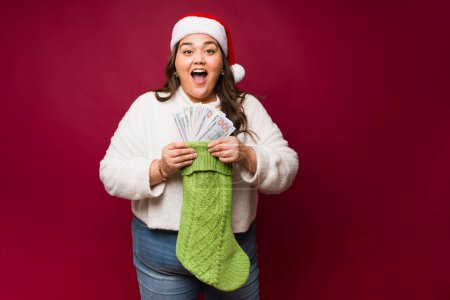 Téléchargez les photos : Joyeux gras femme latine souriant regardant surpris et heureux avec beaucoup d'argent comme un cadeau à l'intérieur de ses bas de Noël - en image libre de droit