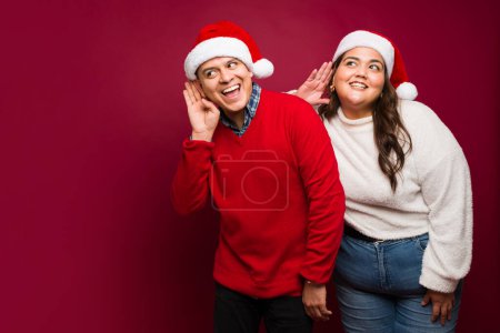 Foto de Hermosa pareja hispana sonriendo mientras intenta escuchar una conversación y espiar sobre los regalos de Navidad para las fiestas - Imagen libre de derechos