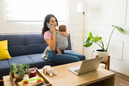 Foto de Atractiva madre cargando a su hijo usando un cabestrillo para bebés hablando por teléfono mientras trabaja desde casa con una computadora portátil en la sala de estar - Imagen libre de derechos
