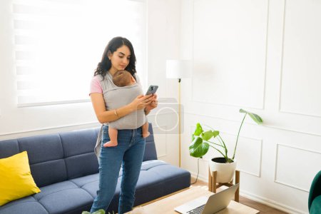 Foto de Madre caucásica en la sala de estar llevando a su hijo recién nacido usando un portabebés mientras envía mensajes de texto en el teléfono inteligente - Imagen libre de derechos