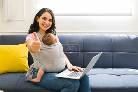 Photo for Positive mom doing a thumbs up feeling happy about doing remote work at home and taking care of her newborn child with a baby carrier - Royalty Free Image