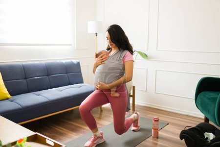 Téléchargez les photos : Fitness maman travailler à la maison tout en portant son nouveau-né dans une fronde bébé regardant exercice heureux - en image libre de droit