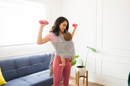 Photo for Strong caucasian mother carrying her newborn child in a baby sling and lifting dumbbell weights and working out at home - Royalty Free Image