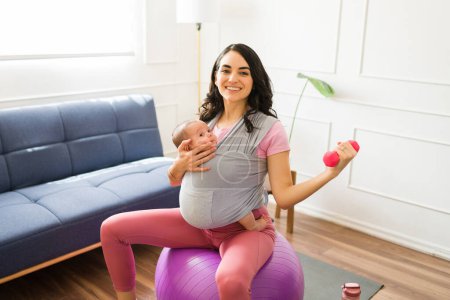 Téléchargez les photos : Portrait d'une maman heureuse en forme souriant portant son nouveau-né dans un bébé fronde exercice à la maison avec haltères et balle de stabilité - en image libre de droit
