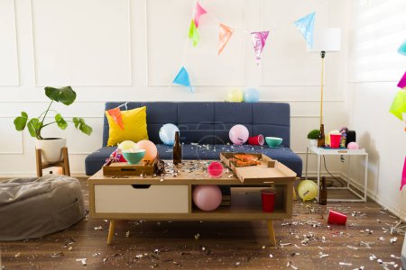 Téléchargez les photos : Maison de salon avec confettis, ballons et décorations de fête ayant un désastre avec des ordures et un désordre après la célébration d'une fête d'anniversaire - en image libre de droit