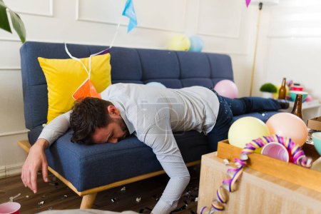 Téléchargez les photos : Homme ivre dans sa trentaine évanoui dormir sur le canapé à la maison avec un désordre après avoir bu beaucoup d'alcool et de bière célébrer une fête - en image libre de droit