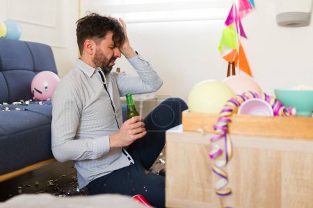 Foto de Hombre borracho molesto con un dolor de cabeza sufriendo una resaca después de celebrar una fiesta de cumpleaños en casa bebiendo cerveza y alcohol - Imagen libre de derechos