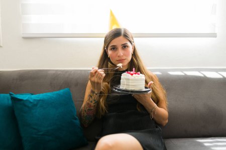Foto de Retrato de una hermosa mujer deprimida comiendo su pastel sola durante su fiesta de cumpleaños sintiéndose sola y triste en casa haciendo contacto visual - Imagen libre de derechos