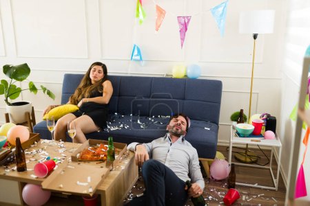Téléchargez les photos : Malade femme hispanique et homme caucasien évanoui ivre et gueule de bois tout en dormant sur le canapé à la suite d'une catastrophe d'une fête d'anniversaire après avoir bu de l'alcool - en image libre de droit