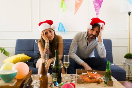 Foto de Atractiva pareja enferma con sombreros de Santa Claus despertando después de una fiesta navideña sufriendo un dolor de cabeza y resaca debido al alcohol - Imagen libre de derechos