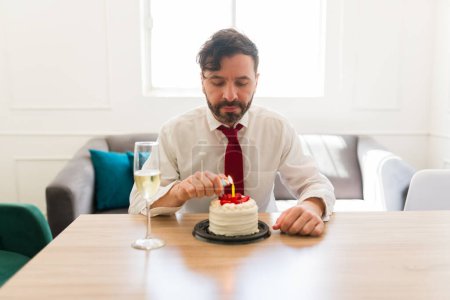 Foto de Triste hombre caucásico encendiendo la vela de su pastel de cumpleaños para hacer un deseo sintiéndose deprimido y solo celebrando en casa - Imagen libre de derechos