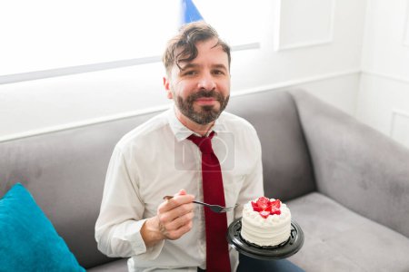 Foto de Feliz hombre caucásico con un sombrero de fiesta sonriendo comiendo delicioso pastel celebrando su cumpleaños relajándose en casa - Imagen libre de derechos