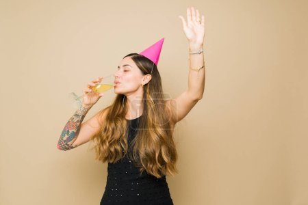 Foto de Mujer joven borracha con un sombrero de fiesta celebrando beber champán y mucho alcohol mientras se divierte mirando feliz - Imagen libre de derechos