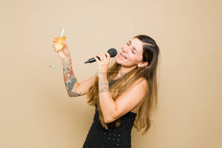 Foto de Mujer joven muy feliz cantando karaoke con un micrófono mientras celebra durante una fiesta y bebe alcohol - Imagen libre de derechos
