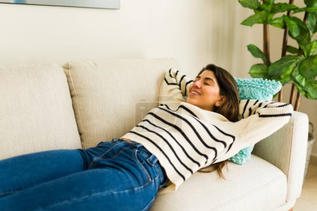 Foto de Feliz joven hispana tumbada en el sofá y sonriendo disfrutando de un acogedor estilo de vida de bienestar durante un tiempo de ocio en la sala de estar - Imagen libre de derechos