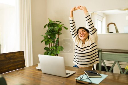 Téléchargez les photos : Heureuse jeune femme détendue étirant le dos et les bras tout en travaillant sur l'ordinateur portable faisant du travail à distance et souriant à la maison - en image libre de droit