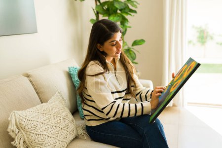 Téléchargez les photos : Jeune femme hispanique détendue avec un style de vie bien-être relaxant sur le canapé et peignant jouissant de son passe-temps artistique dans sa maison confortable - en image libre de droit