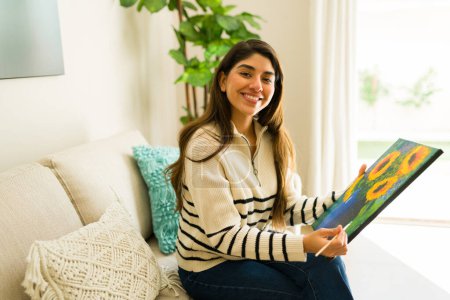 Foto de Alegre joven latina sonriendo haciendo contacto visual mientras relaja la pintura luciendo feliz disfrutando de su hobby artístico en casa - Imagen libre de derechos