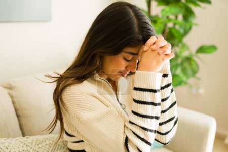 Photo for Spiritual religious hispanic young woman saying a prayer at home and praying for her wellbeing at her cozy home - Royalty Free Image