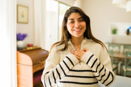 Téléchargez les photos : Spirituel hispanique jeune femme toucher son cœur poitrine tout en se sentant l'amour et détendu sourire à la maison - en image libre de droit