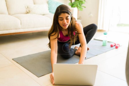 Téléchargez les photos : Latino jeune femme regarder une vidéo de fitness sur l'ordinateur portable tout en exerçant à la maison en appréciant son passe-temps - en image libre de droit