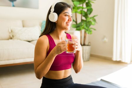Photo for Smiling fit young woman looking happy drinking chamomile tea at home and listening to relaxing music with headphones while practicing yoha - Royalty Free Image