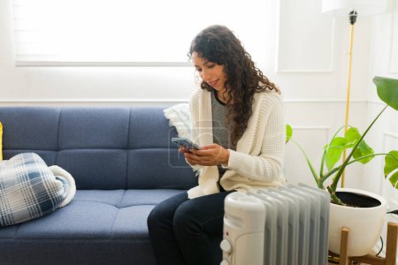 Téléchargez les photos : Joyeux jeune femme avec les cheveux bouclés souriant et textos sur le smartphone tout en se relaxant à la maison assis sur le canapé du salon portant un pull - en image libre de droit