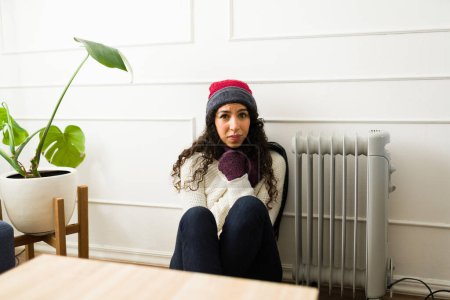 Foto de Hermosa mujer joven molesta sintiendo frío usando ropa de invierno y un sombrero de punto temblando sentado junto al radiador de calefacción en casa durante el clima de otoño - Imagen libre de derechos