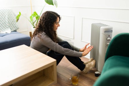 Téléchargez les photos : Heureuse jeune femme détendue réchauffant ses pieds et ses mains dans le radiateur d'huile et souriant à la maison profitant du temps d'hiver - en image libre de droit