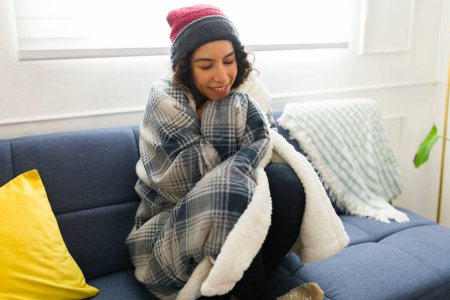 Foto de Relajada hermosa joven con un sombrero de punto sonriendo y disfrutando del clima invernal envuelto en una manta relajándose en el sofá y sonriendo en casa - Imagen libre de derechos