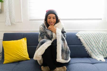 Photo for Smiling beautiful young woman looking happy and relaxed while wrapped in a blanket warming up on the couch while enjoying winter weather - Royalty Free Image