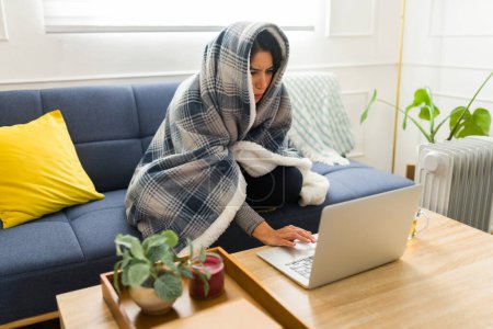 Téléchargez les photos : Jeune femme se sentant très froid sans chauffage à la maison enveloppé dans une couverture assise sur le canapé en utilisant l'ordinateur portable achats en ligne avec le chauffage - en image libre de droit