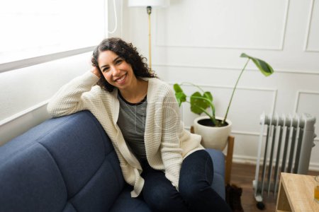 Téléchargez les photos : Belle jeune femme latine souriante tout en se relaxant sur le canapé à la maison et en profitant du réchauffement pendant l'automne regardant heureux - en image libre de droit