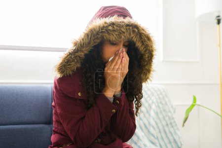 Foto de Mujer joven hispana enferma con una chaqueta sonándose la nariz sintiéndose enferma sufriendo un mal resfriado descansando en casa durante el invierno - Imagen libre de derechos