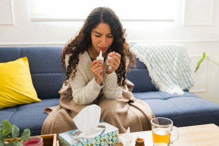 Foto de Mujer joven enferma usando un aerosol nasal mientras está enferma con un resfriado durante el clima invernal y descansando en la sala de estar con té de manzanilla - Imagen libre de derechos