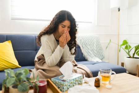 Foto de Mujer joven enferma que sufre un resfriado o la gripe sonándose la nariz con tejidos mientras descansa con una manta en el sofá durante el invierno - Imagen libre de derechos