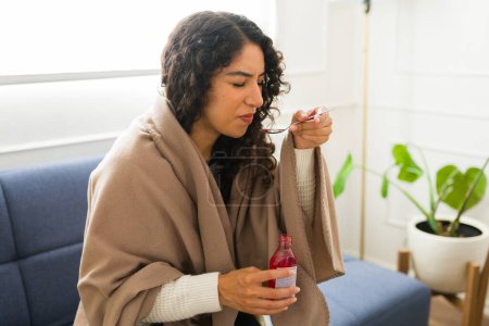 Foto de Malestar latino mujer joven que toma jarabe para la tos y medicamentos mientras sufre un resfriado o gripe y se siente mal en casa envuelto en una manta caliente - Imagen libre de derechos