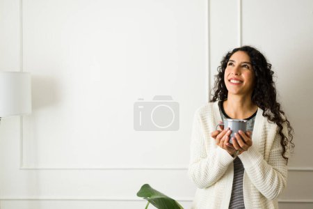 Photo for Thoughtful hispanic young woman smiling and thinking about the cold autumn weather enjoying drinking coffee - Royalty Free Image