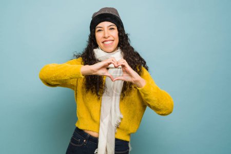 Photo for Cheerful beautiful young woman wearing a knit hat and scarf enjoying the cold weather making a heart love gesture loving the winter - Royalty Free Image