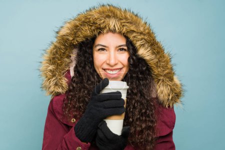 Photo for Hispanic beautiful young woman wearing a big winter jacket smiling looking happy enjoying drinking a coffee to go during cold weather - Royalty Free Image
