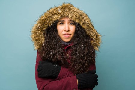 Photo for Portrait of a beautiful hispanic young woman wearing gloves and winter clothing shivering in the cold weather during winter while snowing - Royalty Free Image