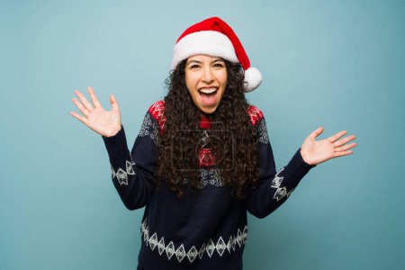 Photo for Excited latin woman screaming looking surprised and happy about the holidays wearing a santa hat and christmas sweater - Royalty Free Image