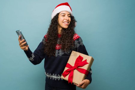 Foto de Atractiva mujer hispana enviando mensajes de texto en el smartphone y celebrando las fiestas recibiendo un regalo de Navidad - Imagen libre de derechos