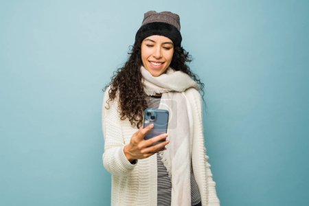 Téléchargez les photos : Belle femme hispanique texter sur le smartphone et sourire en utilisant les médias sociaux portant des vêtements chauds pour le froid de l'hiver - en image libre de droit