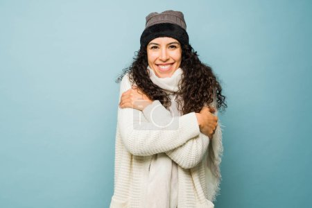 Photo for Cold young woman shivering using a knit hat and scarf enjoying the cold weather during the winter against a studio blue background - Royalty Free Image