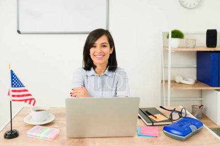 Photo for Cheerful woman teacher smiling looking happy sitting at her desk waiting to start teaching English to students in the classroom - Royalty Free Image