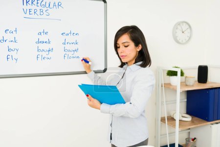 Belle femme travaillant comme professeur d'anglais écrivant des verbes sur le tableau blanc tout en enseignant une langue étrangère
