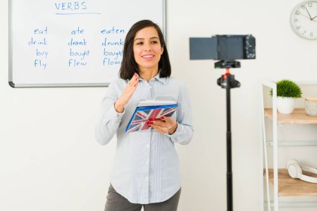 Photo for Beautiful happy virtual teacher filming a video with a camera to do an online streaming English class lessons - Royalty Free Image