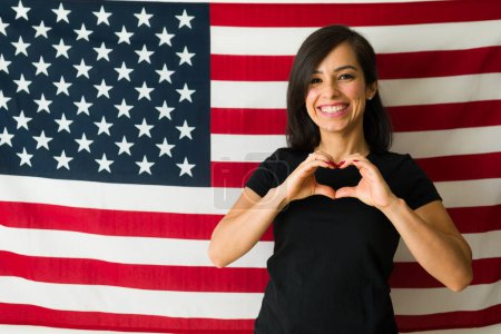 Foto de Patriota atractiva mujer sonriendo haciendo un gesto de corazón amando el sueño americano frente a la bandera de Estados Unidos en el concepto de migración - Imagen libre de derechos