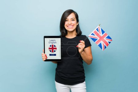 Téléchargez les photos : Belle femme caucasienne tenant le drapeau du Royaume-Uni et montrant une tablette avec une application d'apprentissage ou un site Web pour étudier l'anglais - en image libre de droit